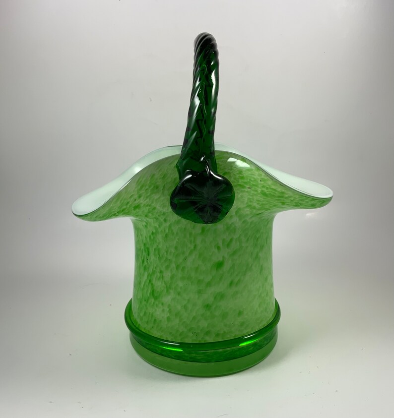 Op de afbeelding: Een groene glazen mand met een witte rand en een gedraaide groene handgreep. De mand heeft een gespikkeld patroon en een effen groene basis. De handgreep is bevestigd aan de mand met een decoratief groen stuk.