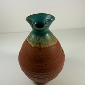 May include: A handmade ceramic vase with a unique shape and color scheme. The vase features a dark teal rim, transitioning to a light green band, and a textured reddish-brown body. The vase has a ruffled top.