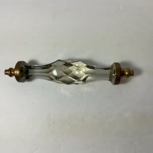May include: A clear glass cabinet pull with a diamond-cut center and brass-colored end caps. The hardware is designed for furniture and has a vintage aesthetic. The pull is approximately 6 inches long.