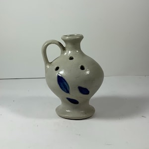 May include: A small, off-white ceramic pitcher with a handle and a rounded body. The pitcher features three blue leaf-shaped designs and several small holes near the top. The base is a circular pedestal.