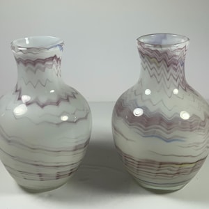 May include: Two white glass vases with a wavy, marbled design in shades of purple, blue, and yellow. The vases have a rounded body and a narrow neck. The design is consistent across both vases, creating a matching set.