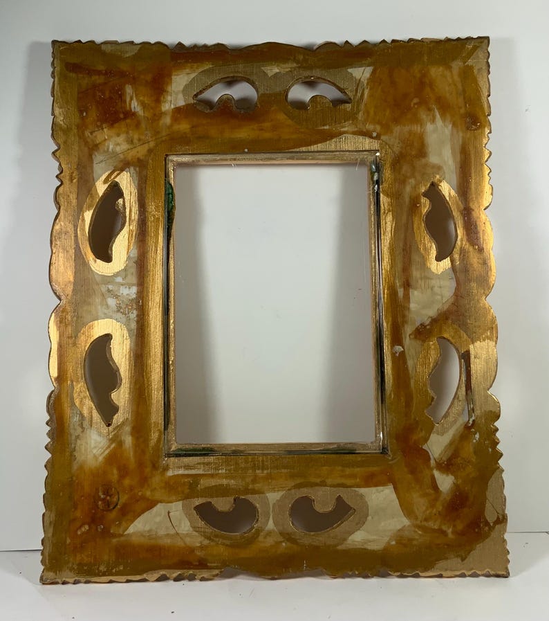 May include: A rectangular picture frame with an ornate design and a distressed gold finish. The frame has decorative cutouts and a scalloped edge. The inner opening is a simple rectangle. The frame has a vintage, antique aesthetic, suitable for a photograph or artwork.