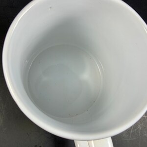 May include: A white ceramic mug with a curved handle. The interior of the mug is also white and appears clean. The mug is empty and sits on a dark surface. The mug is a standard size for coffee or tea.