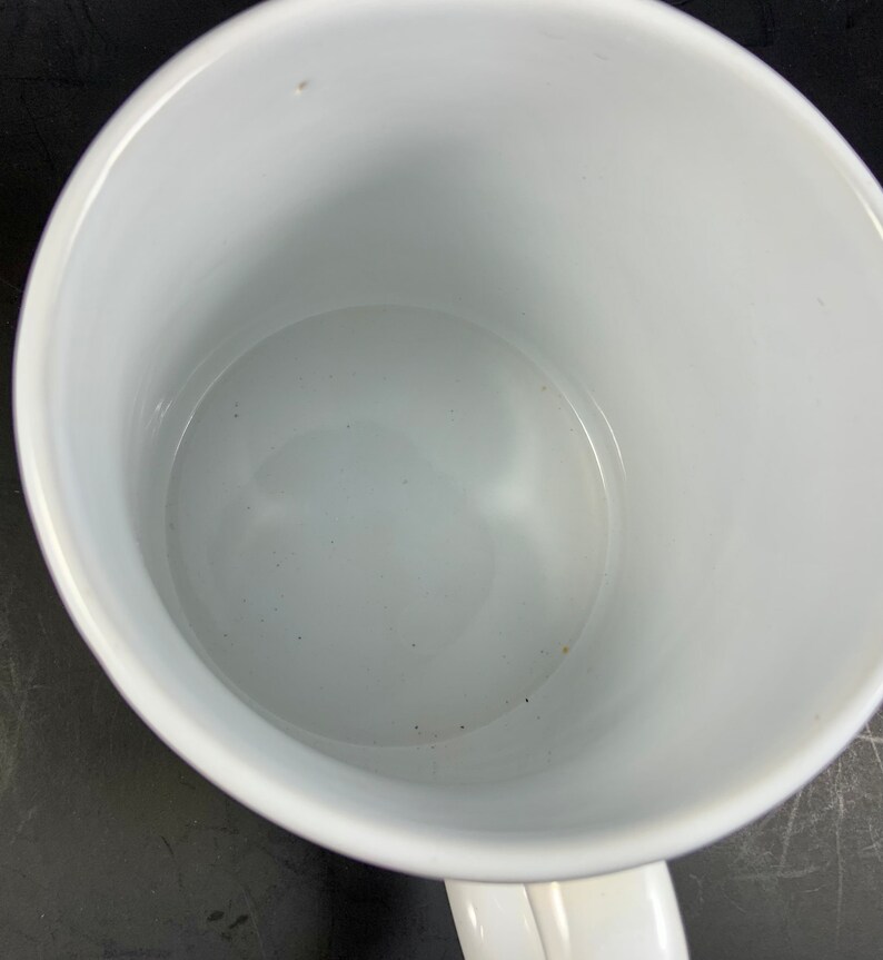 May include: A white ceramic mug with a curved handle. The interior of the mug is also white and appears clean. The mug is empty and sits on a dark surface. The mug is a standard size for coffee or tea.
