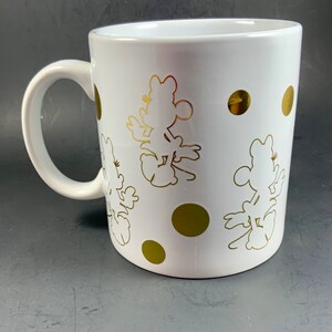 May include: White ceramic mug featuring gold silhouettes of a cartoon character and gold polka dots. The mug has a curved handle and a glossy finish. The design is repeated around the mug.