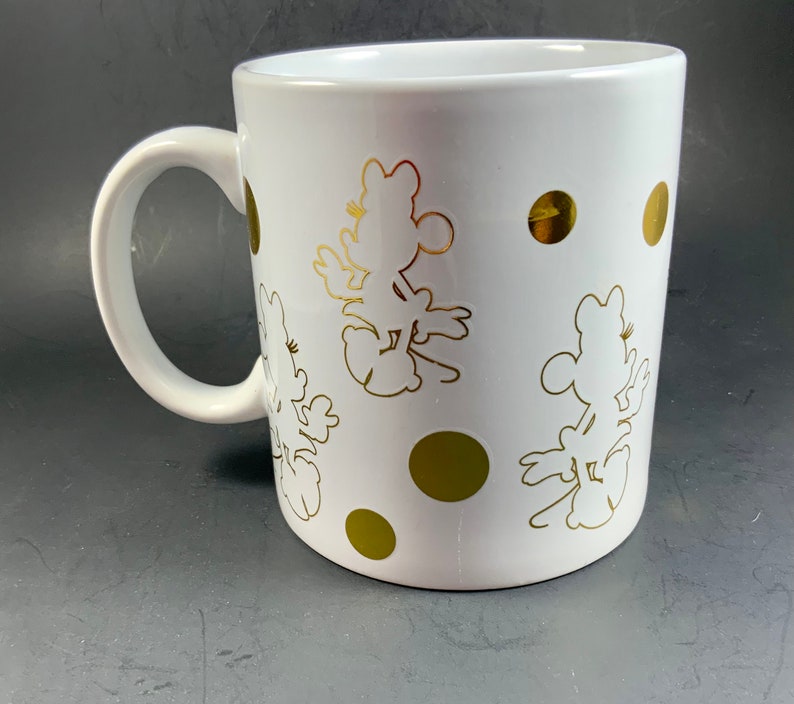 May include: White ceramic mug featuring gold silhouettes of a cartoon character and gold polka dots. The mug has a curved handle and a glossy finish. The design is repeated around the mug.