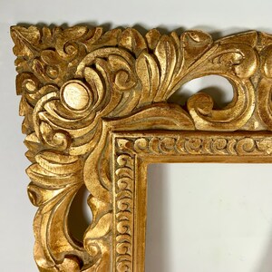 May include: Close-up of a gold-coloured, ornate picture frame. The frame features detailed carvings of swirling leaves and floral patterns. The frame's design is symmetrical, with rounded corners and a rectangular opening.