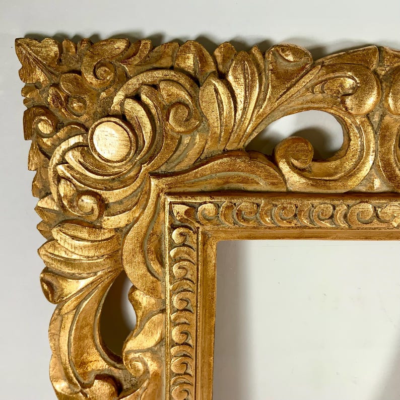 May include: Close-up of a gold-coloured, ornate picture frame. The frame features detailed carvings of swirling leaves and floral patterns. The frame's design is symmetrical, with rounded corners and a rectangular opening.