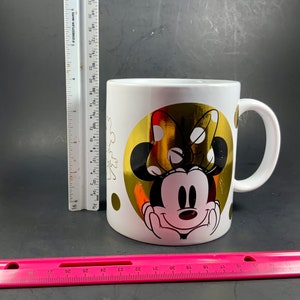 May include: White ceramic mug featuring Minnie Mouse with a gold foil background. The design includes a large gold circle with Minnie's face and bow, and smaller gold polka dots. The mug has a white handle.