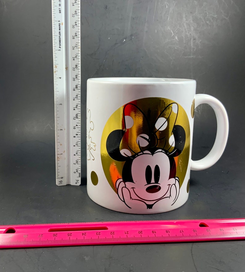 May include: White ceramic mug featuring Minnie Mouse with a gold foil background. The design includes a large gold circle with Minnie's face and bow, and smaller gold polka dots. The mug has a white handle.
