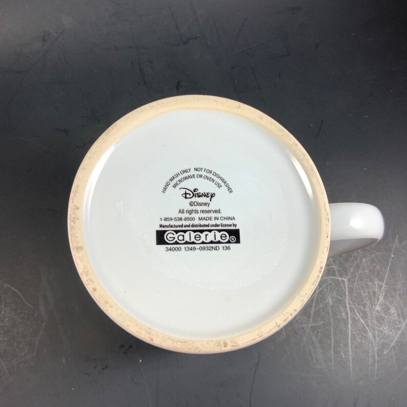 May include: White ceramic mug with a handle, featuring the Disney logo and text. The bottom of the mug displays the Disney copyright information, manufacturing details, and the Galerie brand. The mug is designed for hand washing only and is not suitable for microwave or oven use.