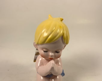 May include: A ceramic figurine of a kneeling cherub with closed eyes and clasped hands. The cherub has blonde hair, pink skin, and small blue accents. The figurine has small, detailed wings.
