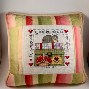 May include: A decorative pillow with a green, pink, and cream striped fabric. The front of the pillow features a cross-stitch design of a cat sitting on a stack of suitcases with the text "In confidence shall be your strength" and "Isaiah 30:15".