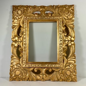 May include: Ornate gold-colored picture frame with detailed carvings. The frame has a rectangular opening and is heavily decorated with floral and scrollwork designs. The frame is made of wood and has a glossy finish, adding to its luxurious appearance.