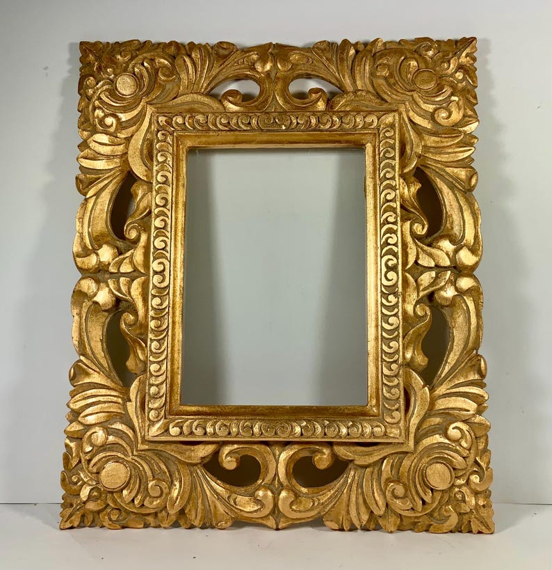 May include: An ornate gold picture frame with detailed carvings. The frame has a rectangular aperture and is heavily decorated with floral and scrollwork designs. The frame is made of wood and has a glossy finish, adding to its luxurious appearance.