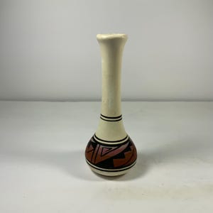 May include: A small, white ceramic vase with a brown and black geometric design. The vase has a narrow neck and a rounded base.