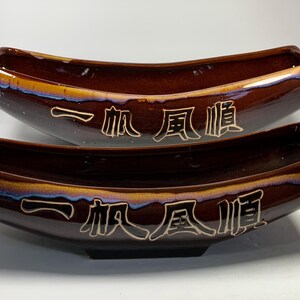 May include: Two brown ceramic bowls with a glossy finish. The bowls are stacked on top of each other. The bowls have a curved shape and are decorated with gold lettering that reads "一帆風順".