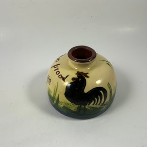 May include: A small, cream-colored ceramic vase with a rooster painted on the front. The rooster is black with a red comb and wattles. The vase has green and blue accents around the rooster and the words "We proud" are written on the side.