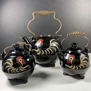 May include: A three-piece black ceramic tea set featuring a teapot, a sugar bowl, and a creamer. Each piece is decorated with a hand-painted rooster motif in red, white, and gold, and includes a gold-toned metal wire handle with a coiled grip.