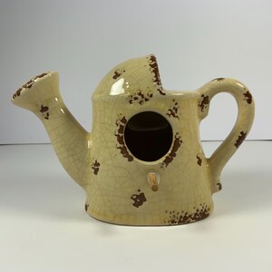 May include: A ceramic birdhouse shaped like a watering can with a distressed, cream-colored finish and brown accents. The birdhouse has a round hole for birds to enter.