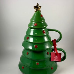 May include: A green ceramic Christmas tree-shaped mug with a gold star on top and red dot ornaments. The mug has a handle and a matching lid. A red tag is attached to the handle.
