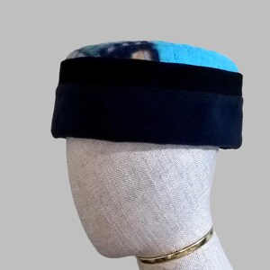 Puede incluir: Un sombrero azul y negro con una banda negra. La parte superior del sombrero tiene un patrón azul y blanco. El sombrero se muestra en una cabeza de maniquí con una pulsera dorada.
