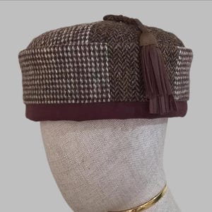 May include: A brown and white patterned hat with a dark brown brim and a tassel. The hat features a houndstooth and herringbone design. The tassel is made of brown leather and is attached to the top of the hat.