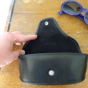 Puede incluir: Un estuche para gafas negro semirr&iacute;gido con cierre de bot&oacute;n a presi&oacute;n y el logotipo de Versace grabado en el forro interior. Junto al estuche abierto hay unas gafas de sol ovaladas de color azul oscuro con lentes oscuras y una marca CE visible.