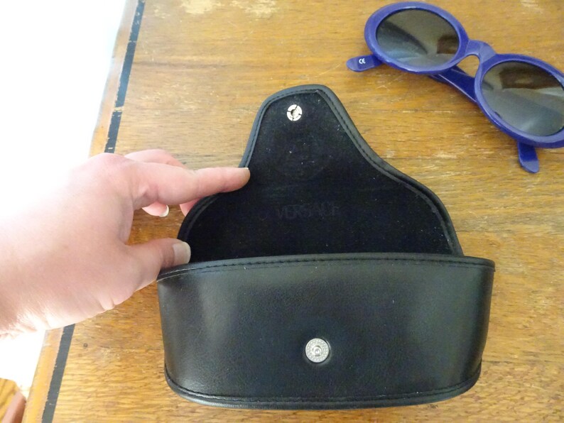 Puede incluir: Un estuche para gafas negro semirr&iacute;gido con cierre de bot&oacute;n a presi&oacute;n y el logotipo de Versace grabado en el forro interior. Junto al estuche abierto hay unas gafas de sol ovaladas de color azul oscuro con lentes oscuras y una marca CE visible.