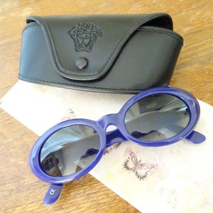 Puede incluir: Gafas de sol ovaladas con montura de pl&aacute;stico azul oscuro brillante y lentes tintadas oscuras, acompa&ntilde;adas de un estuche r&iacute;gido de cuero sint&eacute;tico negro con el logotipo de la cabeza de Medusa en relieve y cierre de bot&oacute;n a presi&oacute;n.