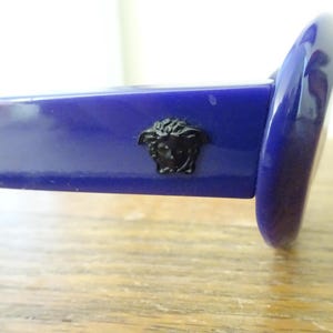 Puede incluir: Un par de gafas de sol azul real brillante con un emblema de cabeza de Medusa negro colocado de forma prominente en la patilla. La montura tiene un acabado liso y pulido y una forma de lente redondeada, mostrando el detalle del logo.