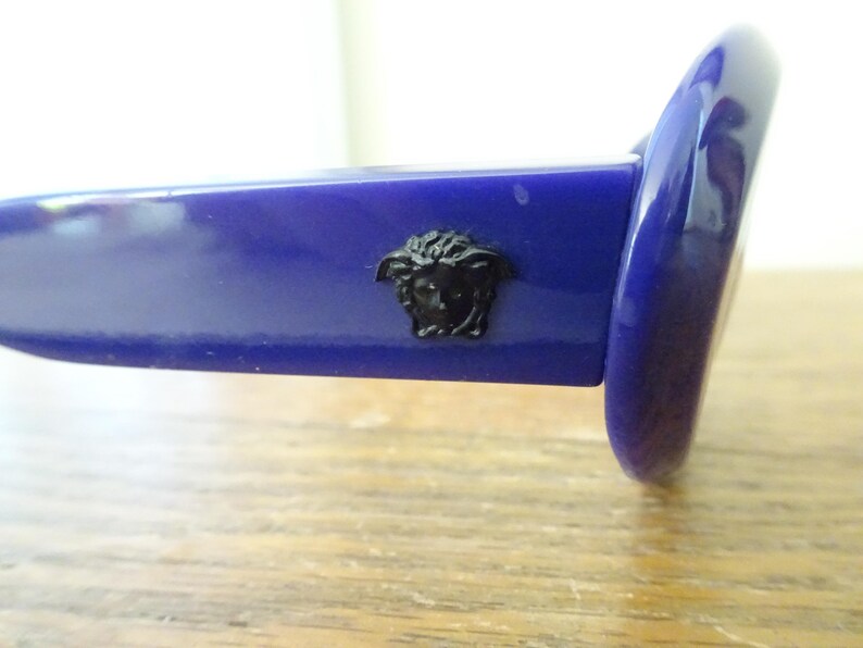 Puede incluir: Un par de gafas de sol azul real brillante con un emblema de cabeza de Medusa negro colocado de forma prominente en la patilla. La montura tiene un acabado liso y pulido y una forma de lente redondeada, mostrando el detalle del logo.