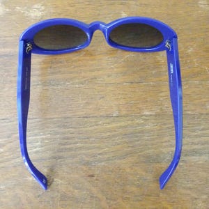 Puede incluir: Un par de gafas de sol ovaladas con montura de pl&aacute;stico azul real brillante y lentes oscuras. El interior de la patilla derecha presenta texto impreso en blanco, mientras que la patilla izquierda muestra marcas adicionales en color blanco.