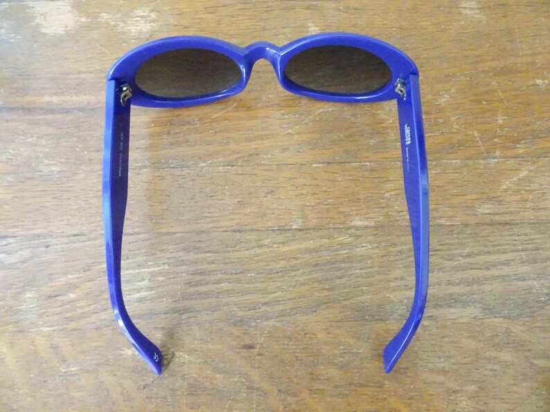 Puede incluir: Un par de gafas de sol ovaladas con montura de pl&aacute;stico azul real brillante y lentes oscuras. El interior de la patilla derecha presenta texto impreso en blanco, mientras que la patilla izquierda muestra marcas adicionales en color blanco.
