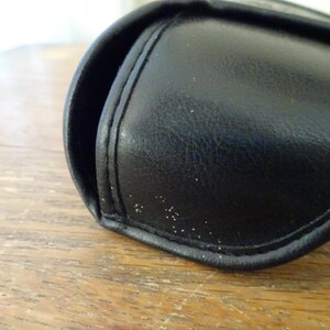 Puede incluir: Un estuche protector negro, semirr&iacute;gido y curvado, con una textura suave similar al cuero y costuras visibles a lo largo de los bordes. El estuche presenta un dise&ntilde;o redondeado con bisagra, una peque&ntilde;a abertura y puntos blancos en la superficie.