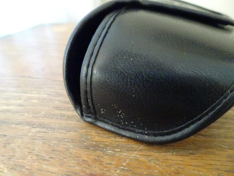 Puede incluir: Un estuche protector negro, semirr&iacute;gido y curvado, con una textura suave similar al cuero y costuras visibles a lo largo de los bordes. El estuche presenta un dise&ntilde;o redondeado con bisagra, una peque&ntilde;a abertura y puntos blancos en la superficie.
