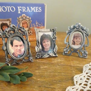 May include: A set of three silver-toned metal photo frames with ornate floral and leaf detailing. Two frames are oval-shaped and one is rectangular. Each frame contains a photograph of a person. The original blue product box is visible behind.