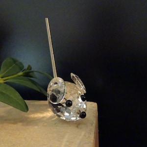 May include: A clear faceted crystal figurine shaped like a mouse with two round ears, two black bead eyes, a small black bead nose, and a long, thin, silver-colored metal spring tail extending vertically from the top of the head.