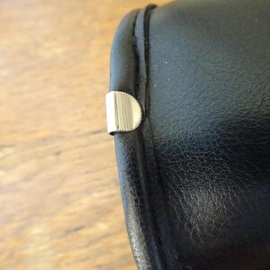 Puede incluir: Un objeto cil&iacute;ndrico de aspecto similar al cuero negro con una superficie oscura y lisa y una costura visible a lo largo del borde. Un peque&ntilde;o clip met&aacute;lico plateado en forma de semic&iacute;rculo con finas estr&iacute;as verticales est&aacute; fijado al borde.
