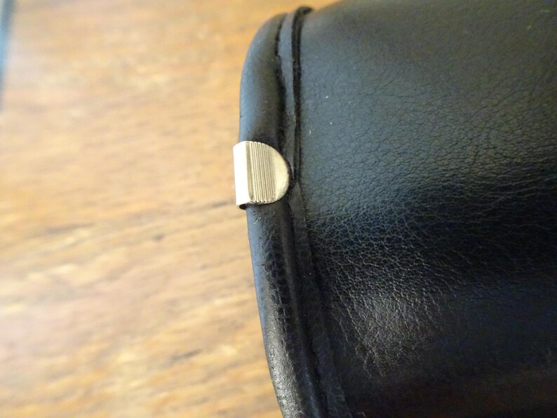 Puede incluir: Un objeto cil&iacute;ndrico de aspecto similar al cuero negro con una superficie oscura y lisa y una costura visible a lo largo del borde. Un peque&ntilde;o clip met&aacute;lico plateado en forma de semic&iacute;rculo con finas estr&iacute;as verticales est&aacute; fijado al borde.