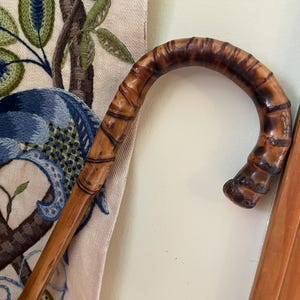 May include: A vintage walking cane with a curved handle. The handle is made of polished bamboo with dark brown and tan tones. The cane is leaning against a wall next to an embroidered fabric with blue and green floral designs.