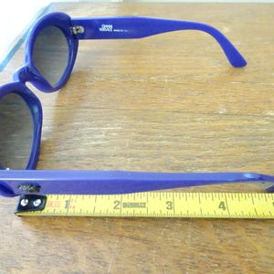 Puede incluir: Gafas de sol de color azul brillante con lentes oscuras y montura gruesa. La patilla interior muestra el texto GIANNI VERSACE MADE IN ITALY. Una cinta m&eacute;trica amarilla se sit&uacute;a bajo la montura, indicando una longitud de unos 14 cent&iacute;metros.