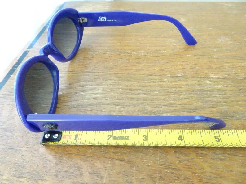Puede incluir: Gafas de sol de color azul brillante con lentes oscuras y montura gruesa. La patilla interior muestra el texto GIANNI VERSACE MADE IN ITALY. Una cinta m&eacute;trica amarilla se sit&uacute;a bajo la montura, indicando una longitud de unos 14 cent&iacute;metros.