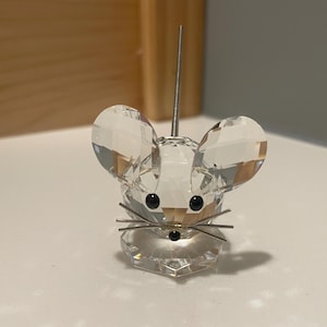 May include: A clear crystal mouse figurine with faceted details. The mouse has large, rounded ears, small black eyes, and metal whiskers. A thin, silver antenna extends from the top of its head. The figurine is displayed on a white surface.