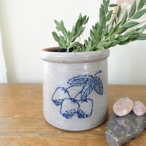 May include: A stoneware crock with a blue strawberry design, filled with green leafy branches. The crock is a light beige color with a darker rim. Decorative stones are in front of the crock on a wooden surface.