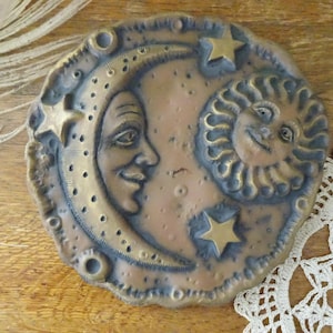 May include: A bronze-colored, circular decorative piece featuring a smiling crescent moon and a sun with a face. Gold-colored stars are placed around the moon and sun. The piece has a textured surface and is likely made of metal.