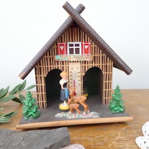May include: A wooden chalet-style thermometer with a brown roof and walls. The chalet features a small window with red shutters and a decorative trim. Miniature figurines of a woman and deer are in front of the thermometer.