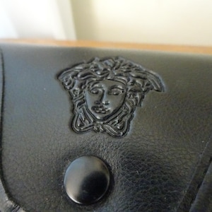 Puede incluir: Un accesorio de cuero negro que presenta un emblema de cabeza de Medusa en relieve centrado en la solapa frontal. La superficie tiene una textura suave con costuras visibles a lo largo de los bordes y un bot&oacute;n de presi&oacute;n circular negro debajo.