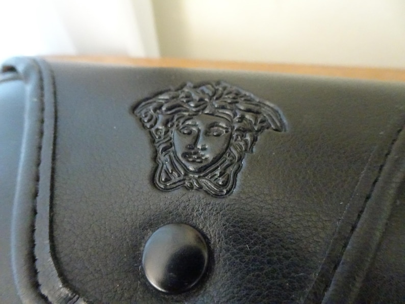 Puede incluir: Un accesorio de cuero negro que presenta un emblema de cabeza de Medusa en relieve centrado en la solapa frontal. La superficie tiene una textura suave con costuras visibles a lo largo de los bordes y un bot&oacute;n de presi&oacute;n circular negro debajo.