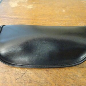 Puede incluir: Un estuche para gafas negro de forma semiovalada con una superficie lisa y mate y bordes cosidos. El estuche presenta un cierre de solapa curvado con un peque&ntilde;o detalle met&aacute;lico de color plateado visible en el lado izquierdo del marco.